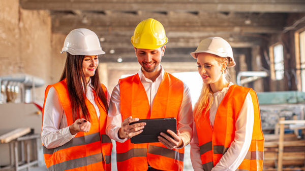 Three young people dressed in orange hi-vis vests and hard hats look at an iPad on a construction site