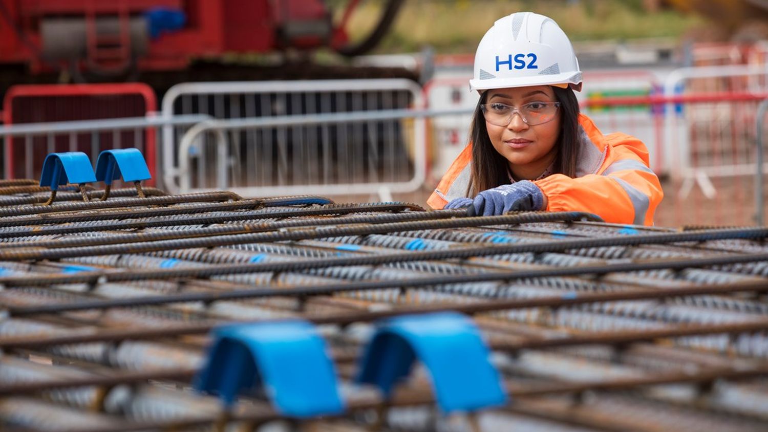 A female HS2 worker in orange hi-vis, grey gloves and white hard hat inspects steel reinforcement bars