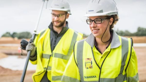 A bearded male Skanska worker wearing hi-vis and a hard hat to the left in the background, with a female skanska worker wearing glasses, a hard hat and hi-vis to the right in the foreground.