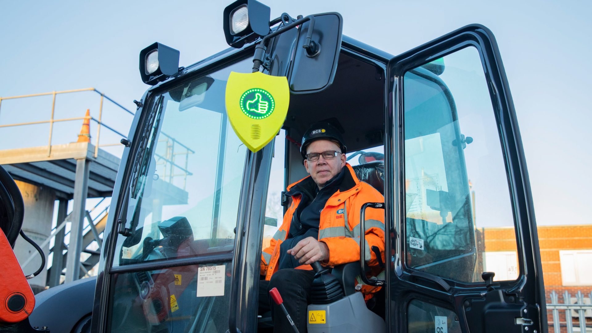 A man in an excavator with an orange high-vis jacket. The excavator has a shield outside with a LED thumbs up.