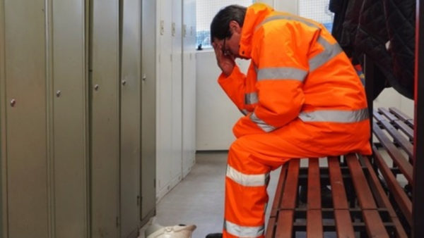 A man in an orange high-vis suit holding his head on one arm, looking sad. There's a hard hat on the floor.
