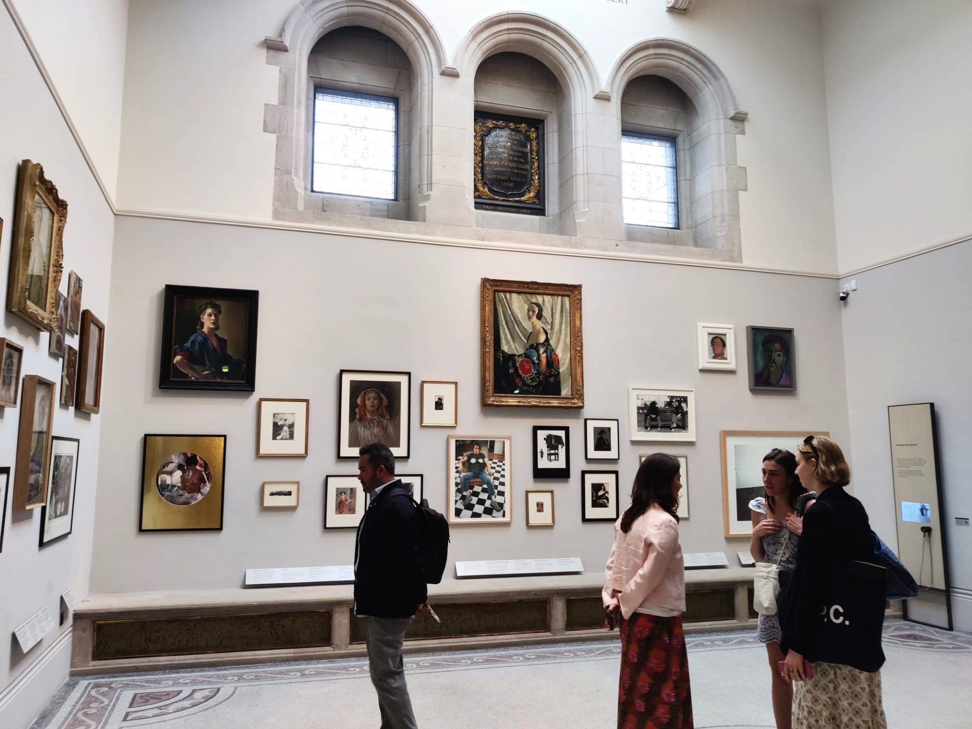 View of the inside of an art gallery with paintings on the walls and people walking around them.