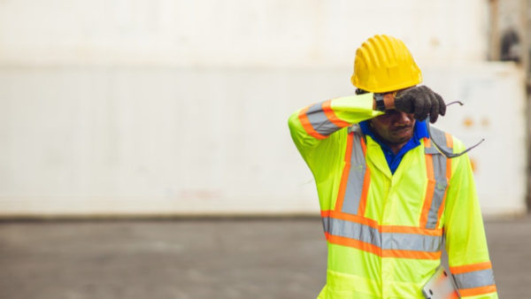 A tired worker in a high-vis jacket and hard hat, rubbing off sweat from his forehead.