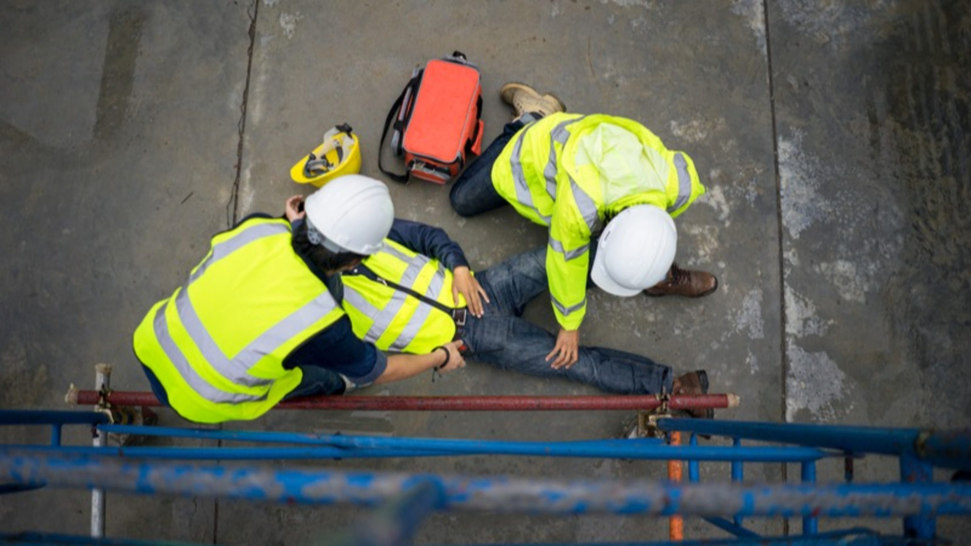 Two workers assisting a third one who is on the floor injured. They are all wearing high-vis jackets.
