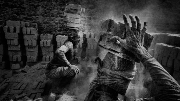 A black and white photo of two people carrying bricks on their heads.