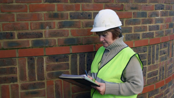 A female building inspector with a white hard hat and a high-vis jacket looking at a clipboard.