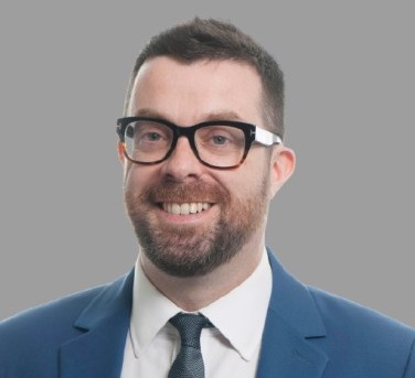 Mugshot of a man with a beard smiling at the camera wearing glasses, a blue jacket, white shirt and a tie.