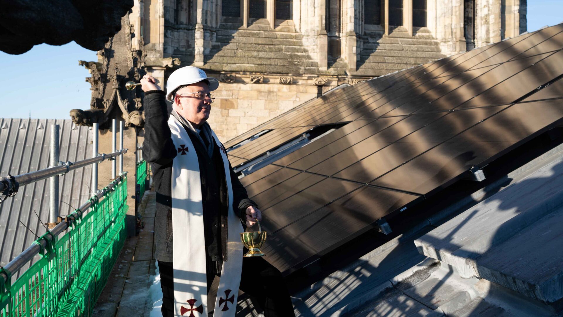 A male priest wearing a white hard hat splashing holy water on solar panels on a roof - York Minster had its new solar panels blessed by the Dean of York.