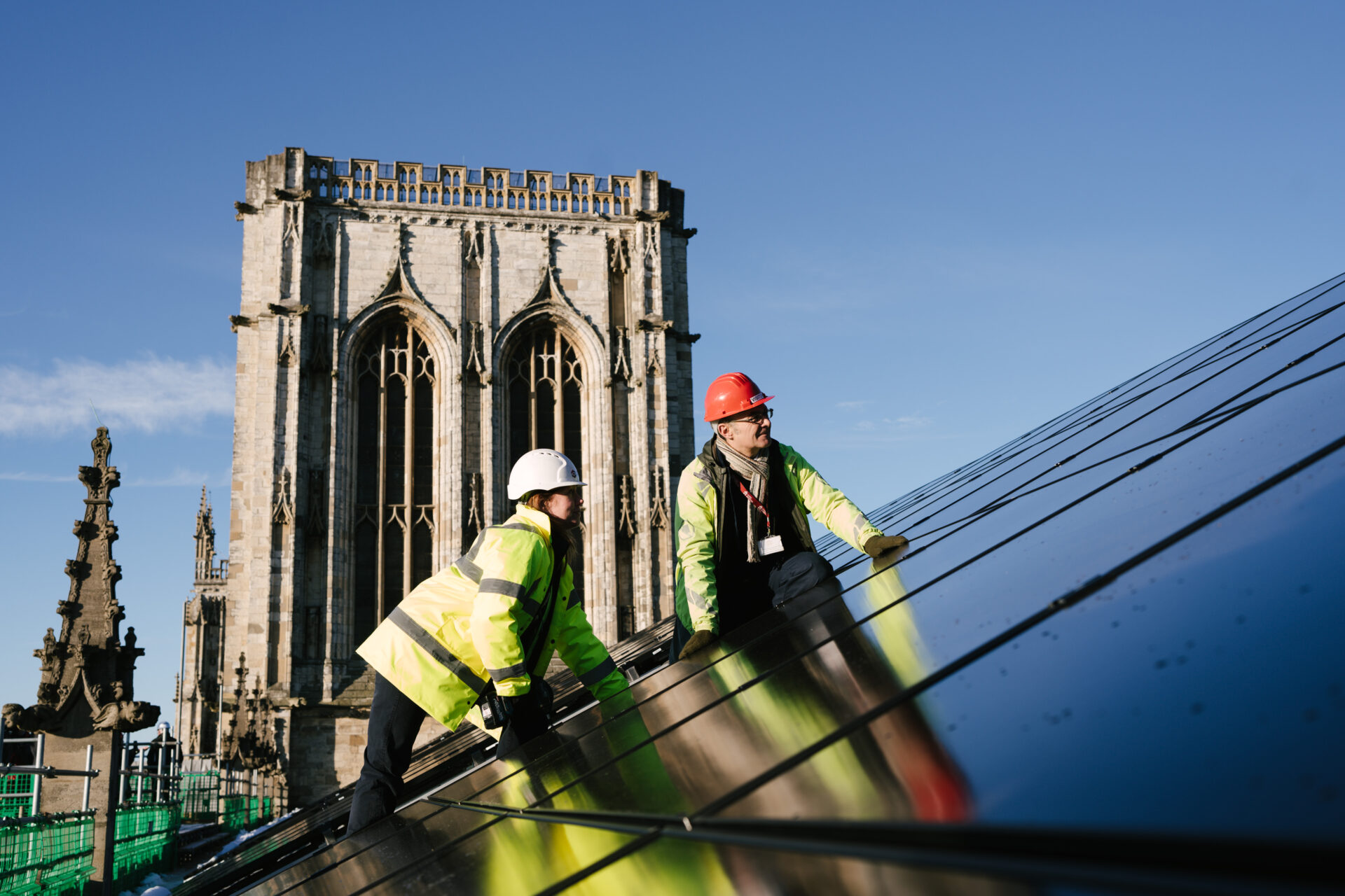 Two workers wearing PPE installing a solar panel on a roof with a cathedral tower on the background - York Minster had its new solar panels blessed by the Dean of York.