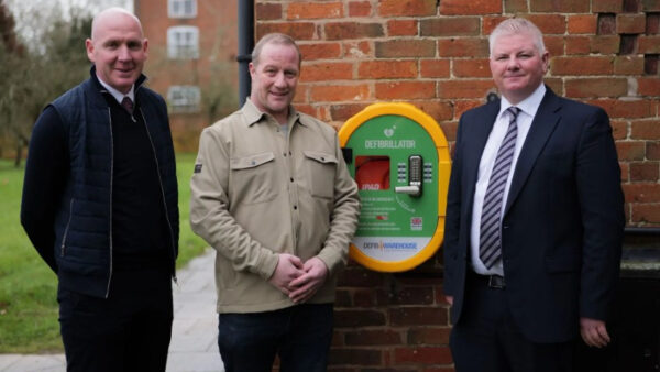 Three men next to a defibrillator installed on a brick wall - Defibrillator saves McCoy Machine Operator's Life