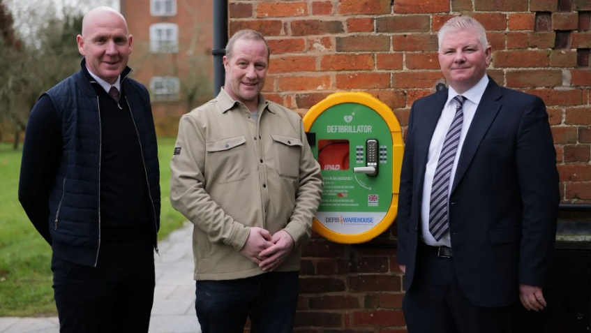 defibrillator Wates - Three men next to a defibrillator installed on a brick wall - Defibrillator saves McCoy Machine Operator's Life