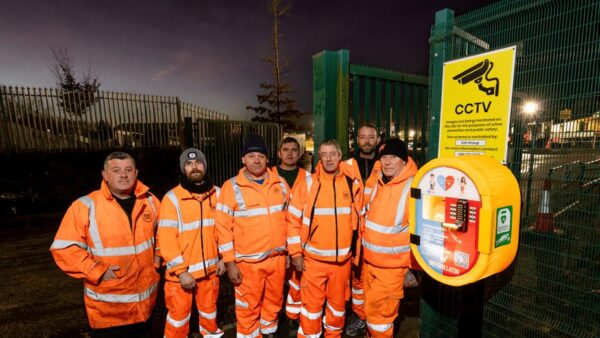 A group of workers wearing orange high-res clothes next to a defibrillator - Esh Group has installed 55 of these machines across its sites