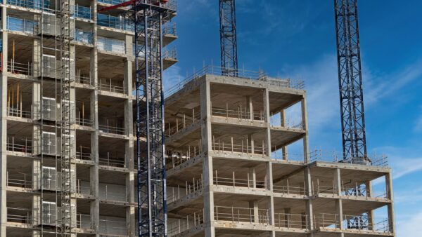 High-Rise building construction site with cranes and scaffolding. Two adjacent high-rise buildings under construction are visible against a clear blue sky. Adrian Gladstone writes that the Building Safety Regulator will need more resources to deal with the delays of the gateways process.